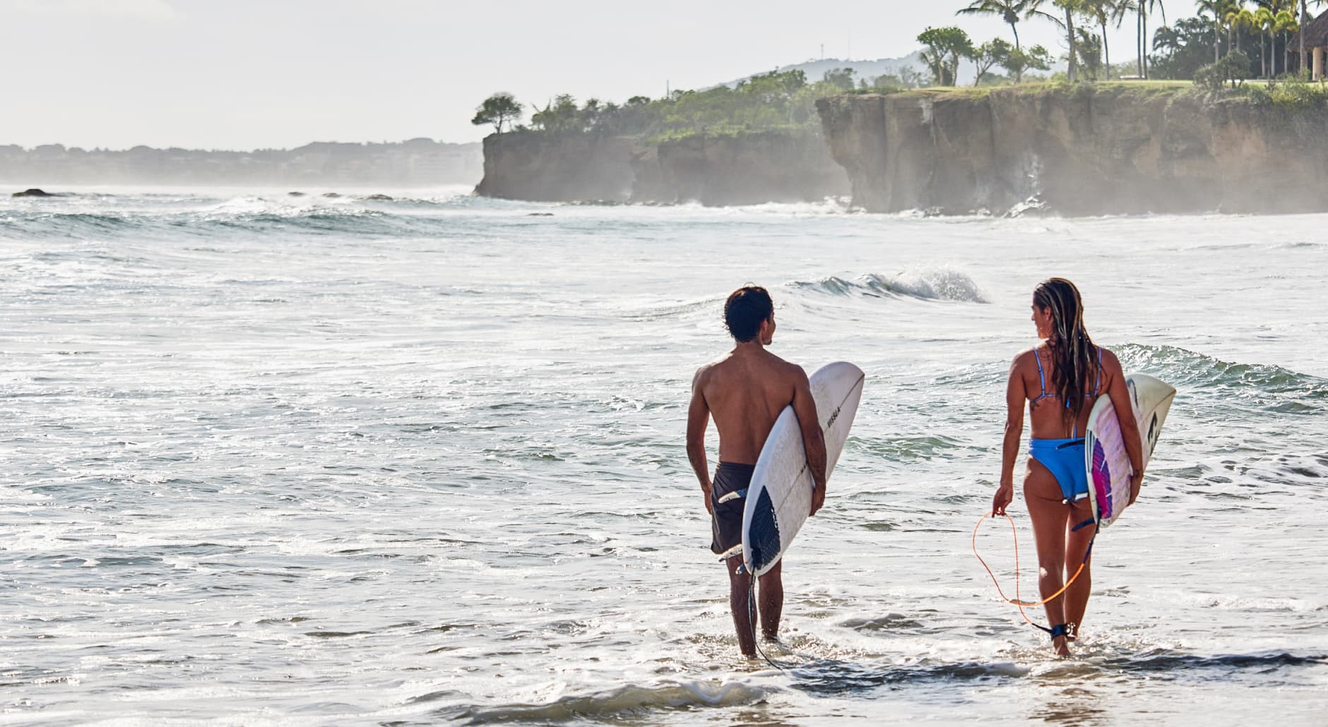 Two people walk along beach with surfboards
